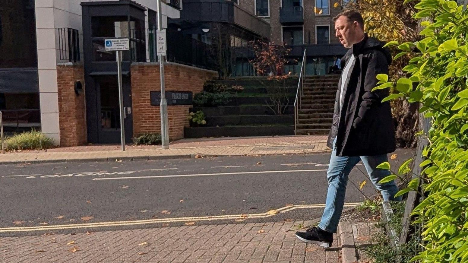 Gregory Assemakis: A- side-on view of a dark-haired man walking on a pavement past a green hedge. He is wearing black shoes, blue jeans, a pale round-necked top and had his hands in the pocket of a black  padded anorak. In the background is a building and two sets of steps.