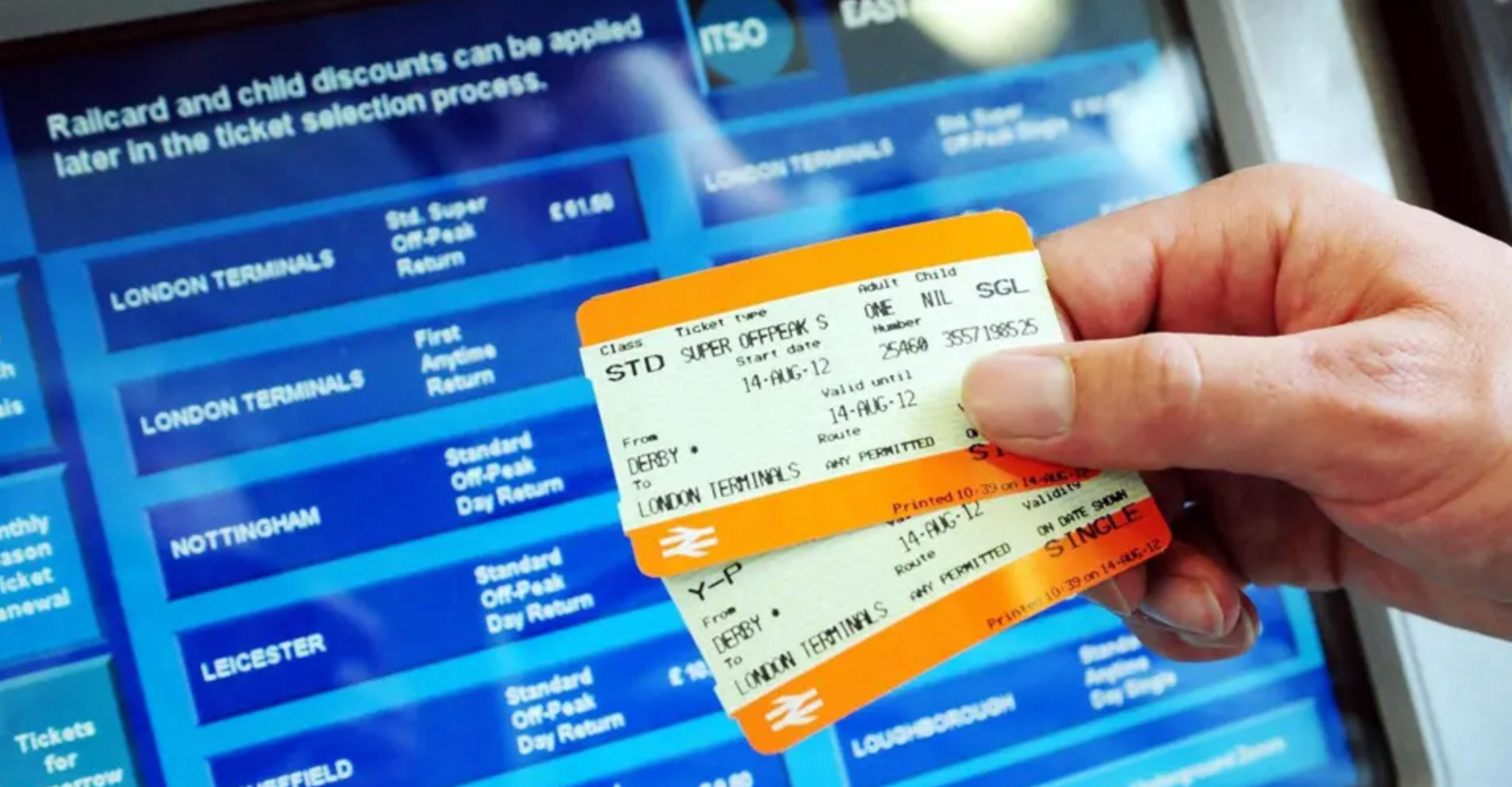 Two rail tickets being held in front of a ticket machine screen with various travel options and ticket types on display.