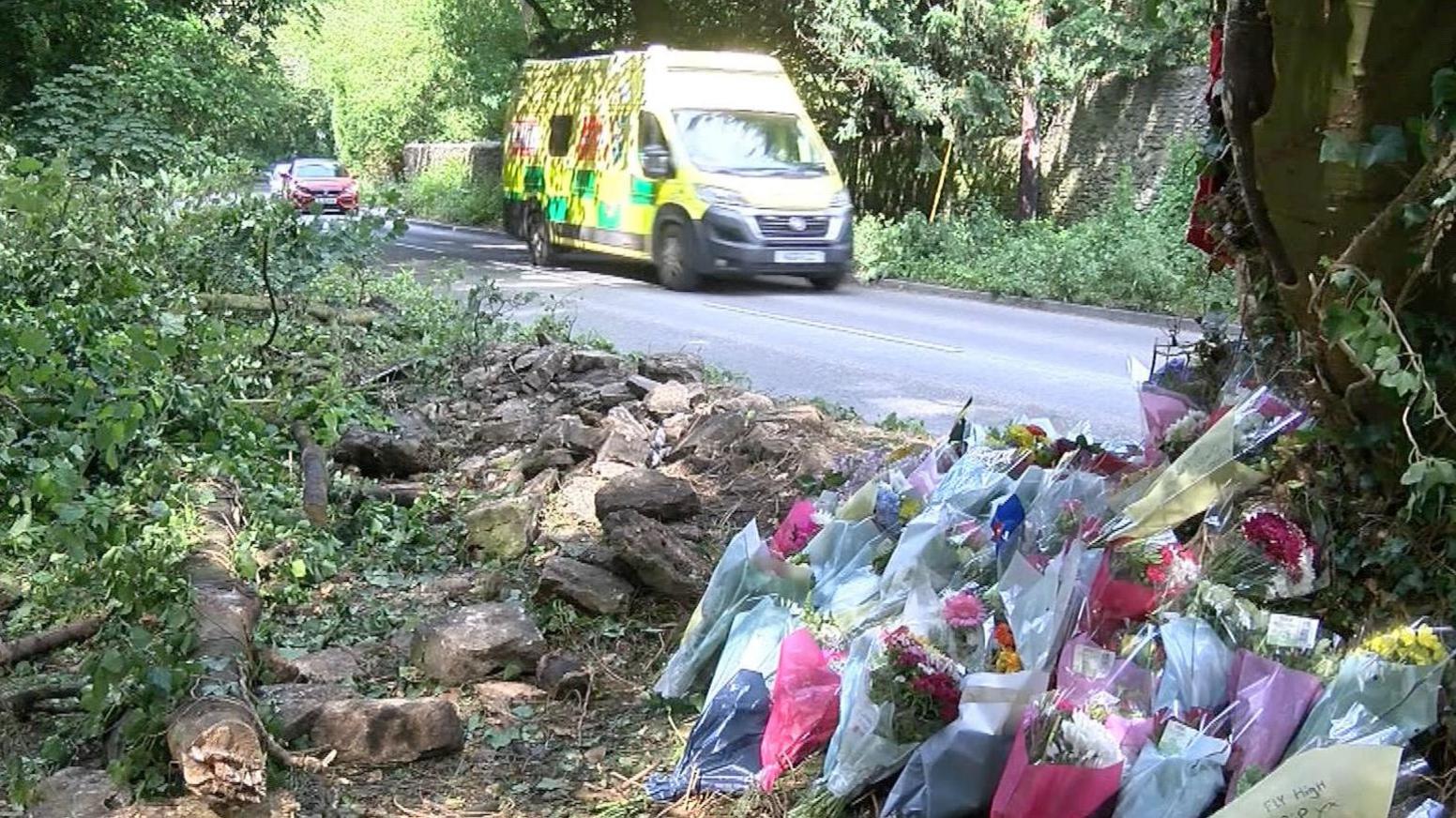 Floral tributes lying against a tree at the scene of the crash in Marcham, with an ambulance and cars driving past in the back ground