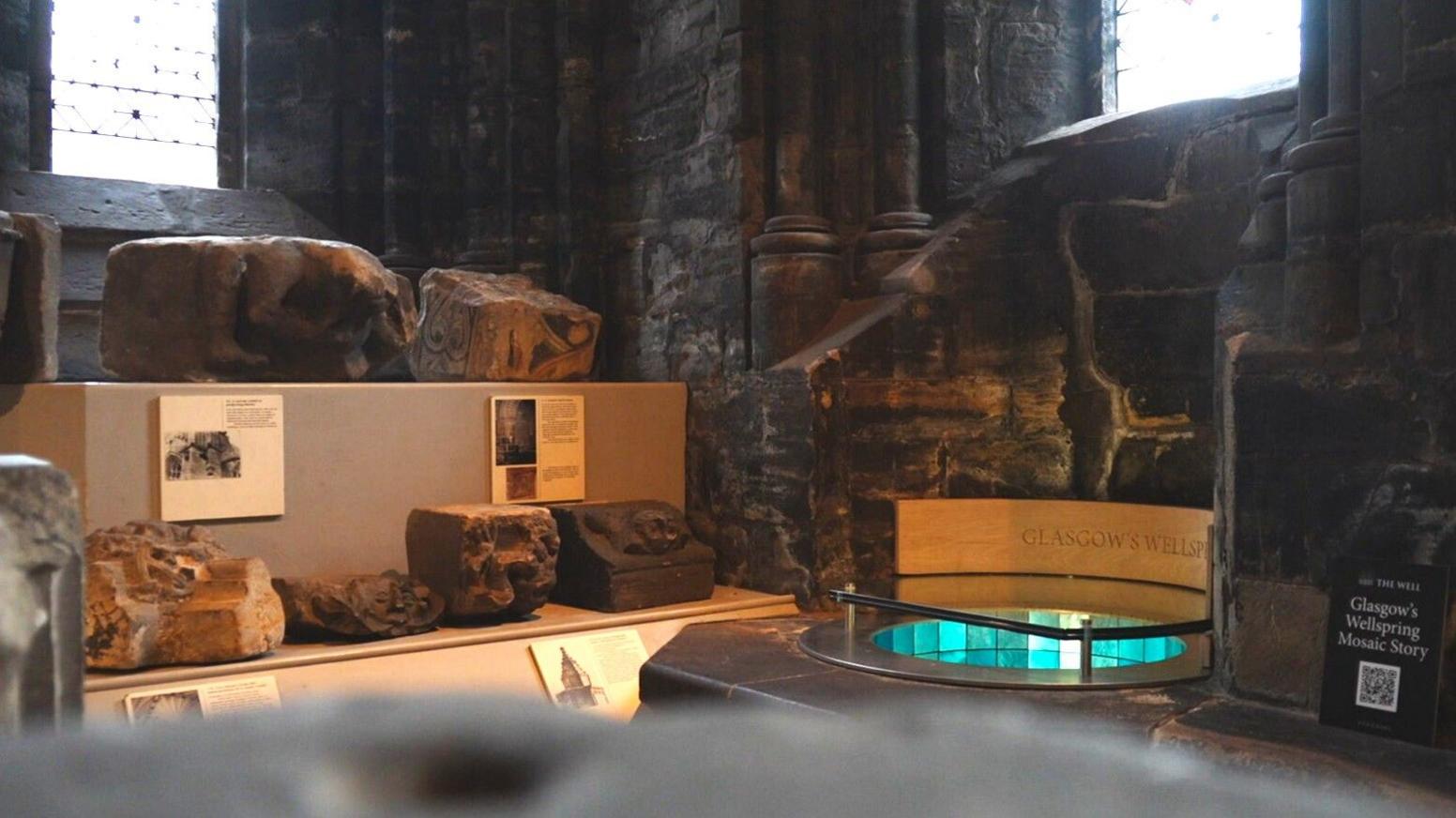 A view from further away of the well in the crypt of Glasgow Cathedral. The mosaic chines up from the well shaft and stained glass windows stand above. The well is partially covered in protective glass and is surrounded by stone.
