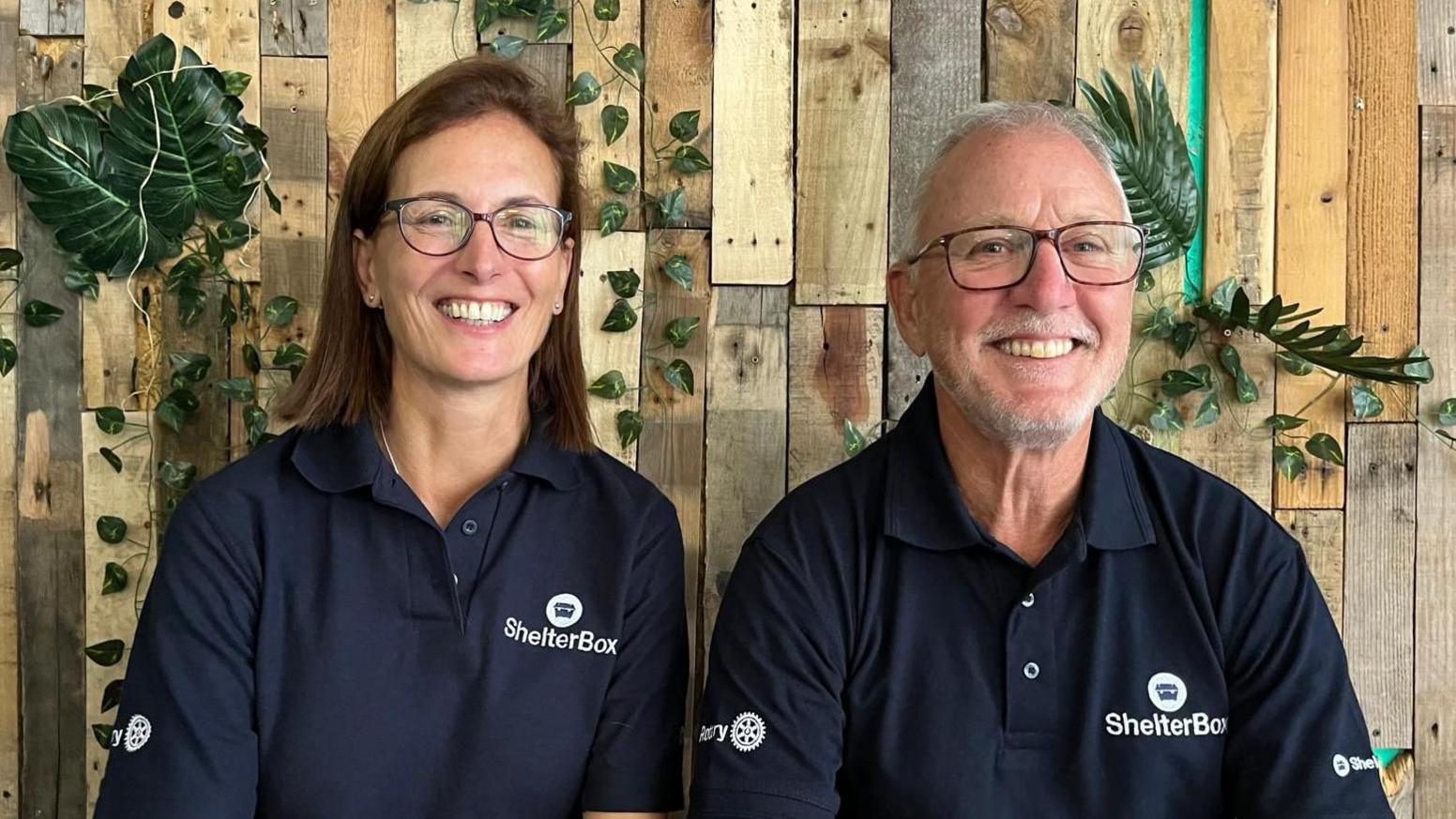 A smiling woman with long brown hair sits beside a man with very short white hair. They are both wearing dark blue polo tops with the ShelterBox logo on them. They are in front of a background of wooden planks and green foliage.