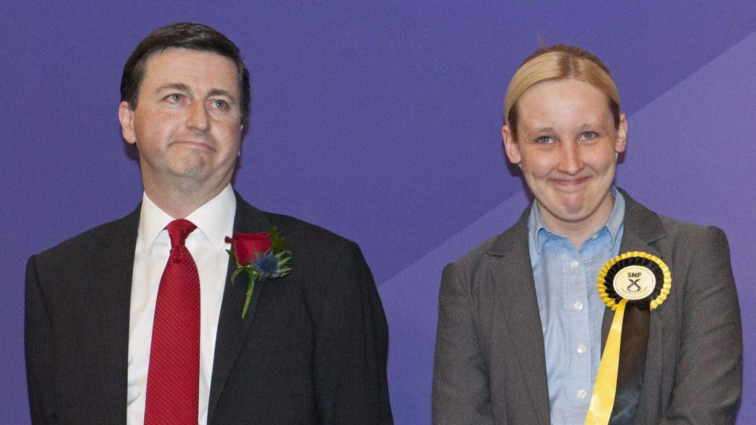 The election count in 2015 - Douglas Alexander -  a dark haired man with a suit, red tie and red rosette, stands next to Mhairi Black, who has  blonde hair and is wearing a grey suit and an SNP rosette