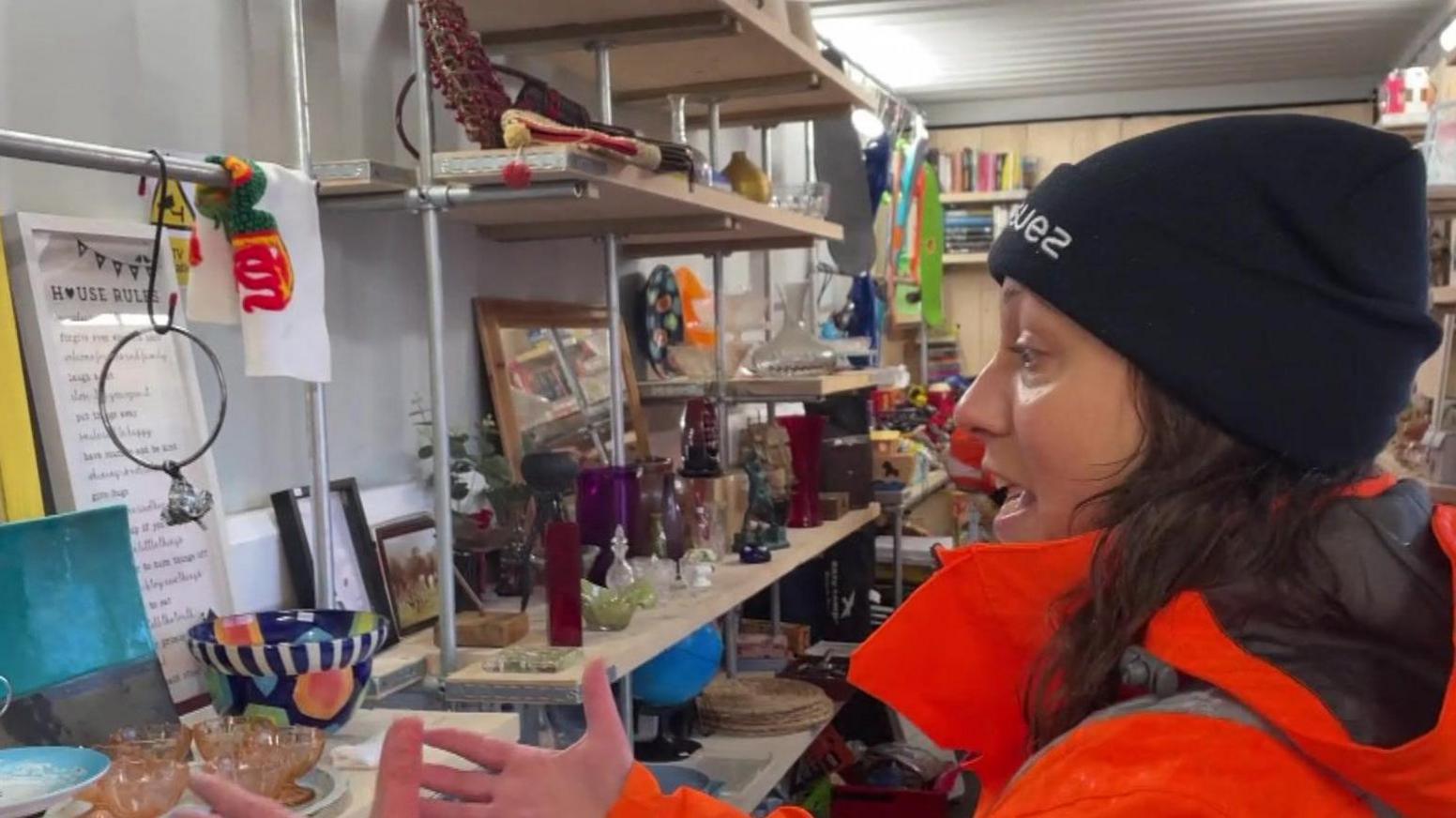 Woman in orange high-vis jacket and black beanie hat with the word Suez on it inside second hand shop at recycling centre. Glass and ceramic items and a mirror are placed on rows of scaffold board shelves.