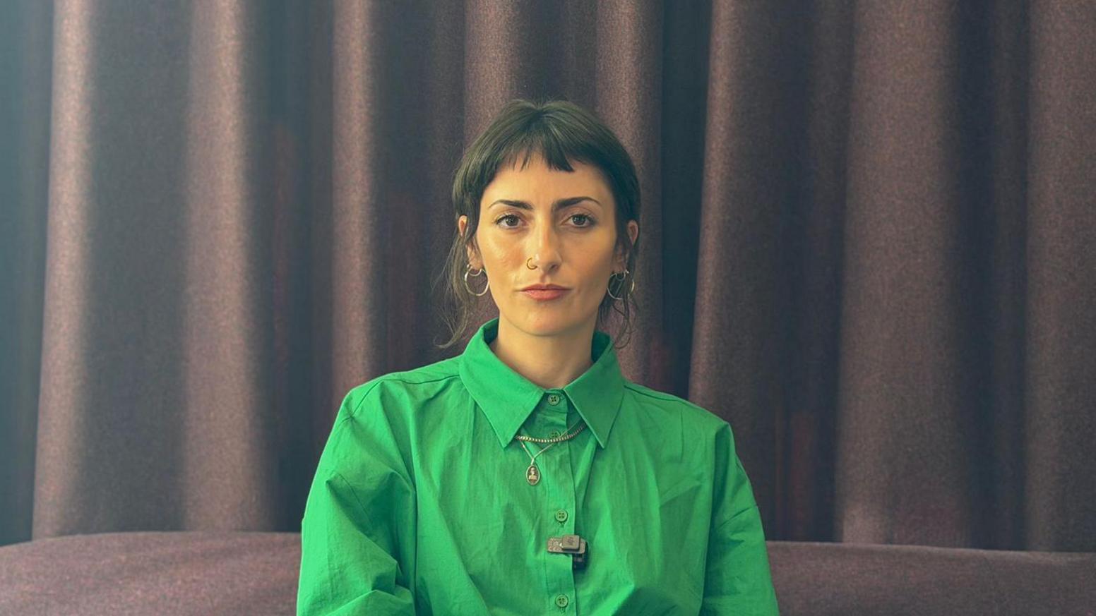 A woman with dark hair is sitting on a brown sofa in front of a brown curtain. She is wearing a green buttoned up shirt and is smiling at the camera