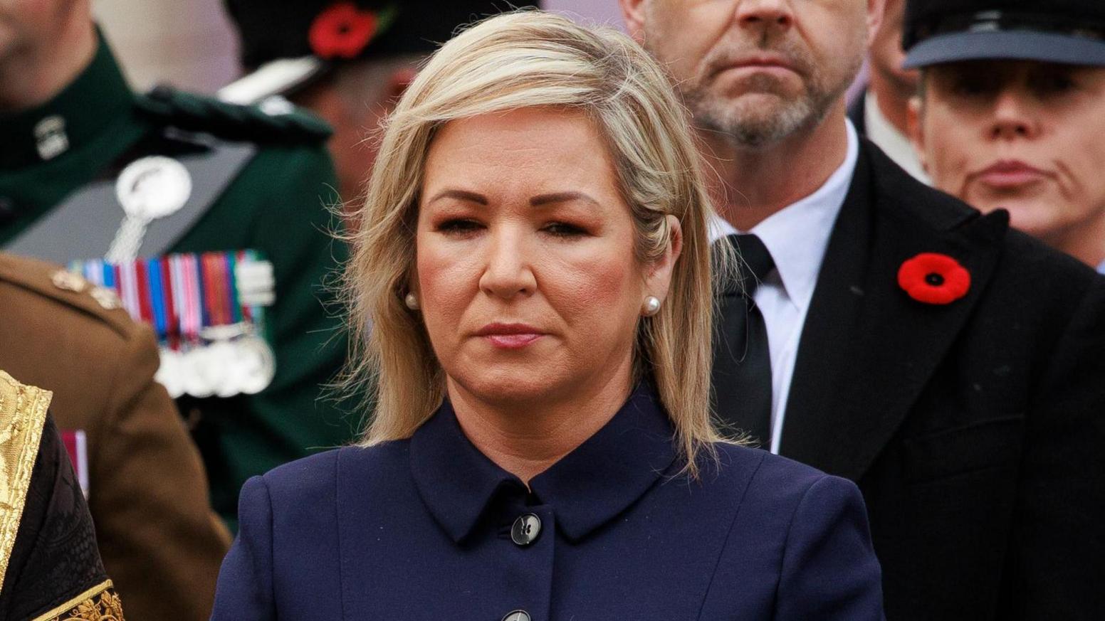 Michelle O'Neill looking solemn in navy suit jacket at remembrance event, men pictured in background are wearing dark clothes, war medals and a poppy