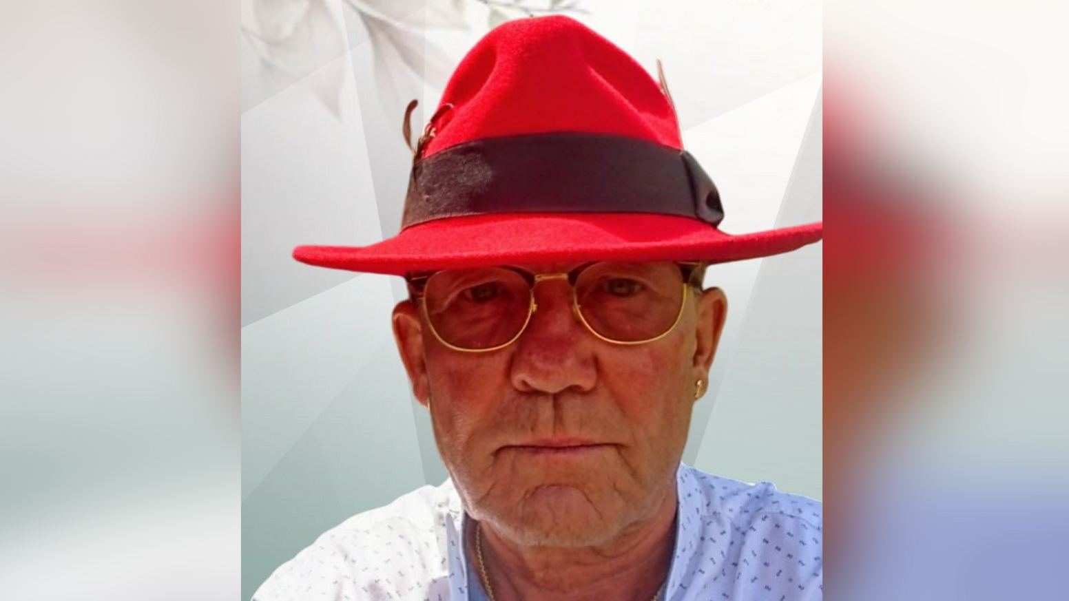 Elderly man with glasses wearing a distinctive red trilby hat. He is staring directly at the camera.