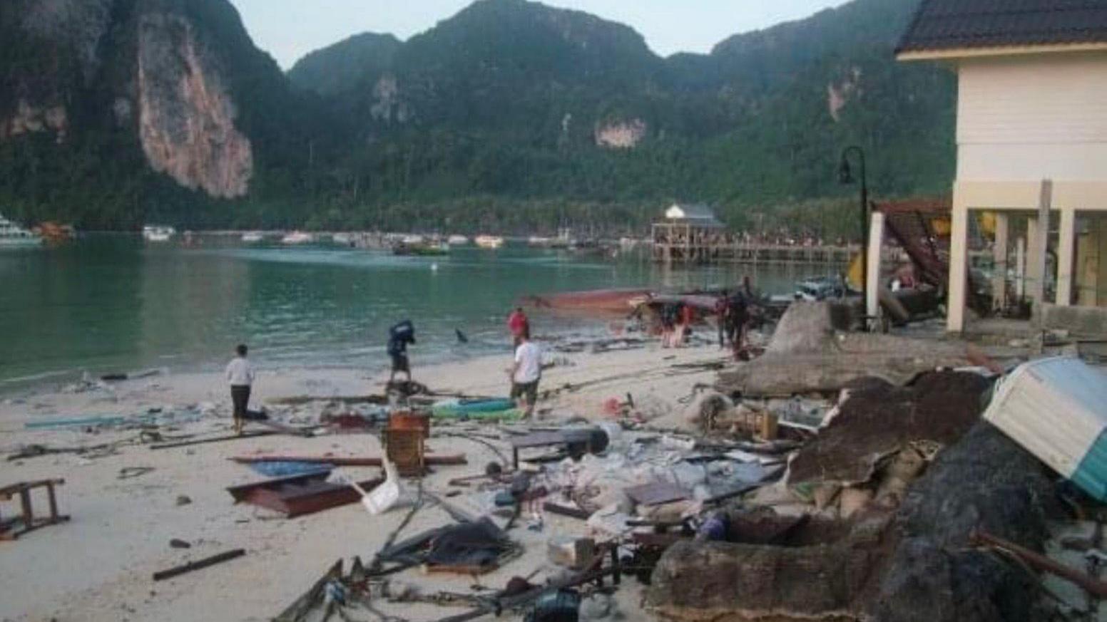 The small harbour of Koh Phi Phi following the tsunami. There is lots of unidentifiable debris and litter scattered across the sand. There are people walking across the beach and assessing the damage. There is a small white beachfront building on stilts to the right which is still standing. In the distance the beach curves around and there is a small wooden pier. In the background there is a large green mountain range.