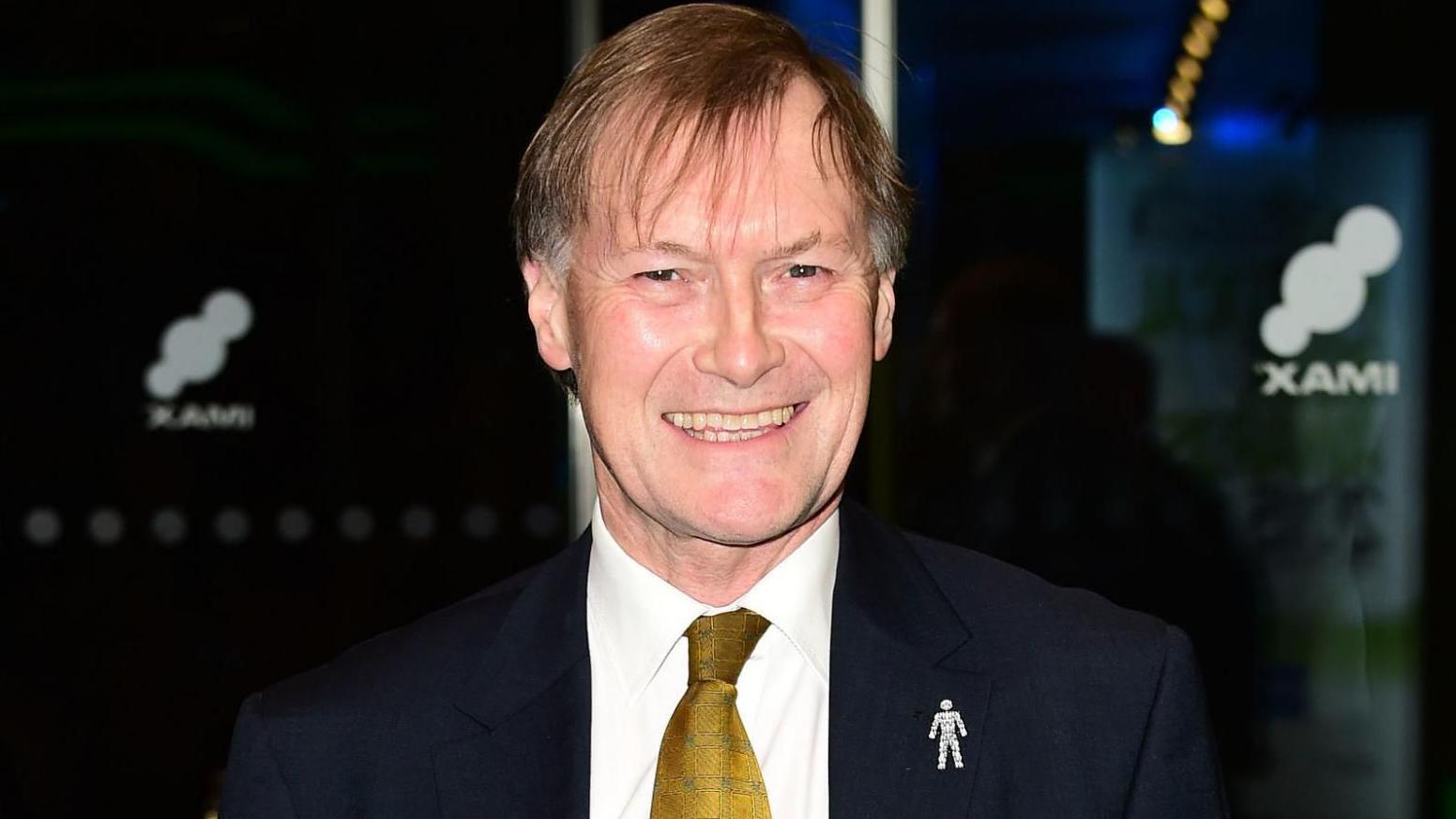 Sir David Amess, wearing a blue suit with a gold patterned tie, stands in front of a dark background with glass doors in the background.