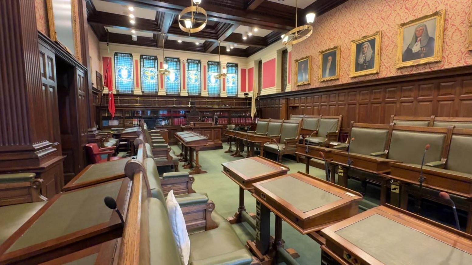 The House of Keys chamber is empty, and has two tiers of seating on both the left and right hand side of the room. The seats are upholstered in green leather and are trimmed with dark wood. The room is panelled in oak and the back wall has six stained glass windows. The wallpapered walls include portraits of former members.