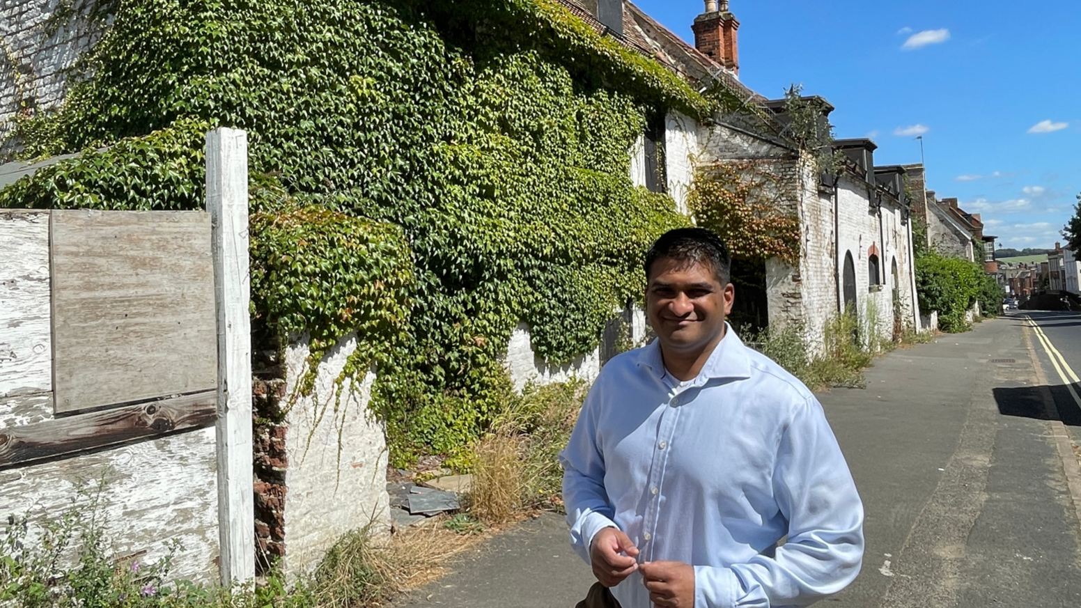 A man wearing a blue shirt standing in front of a derelict building with green leaves growing out of it in Newmarket.