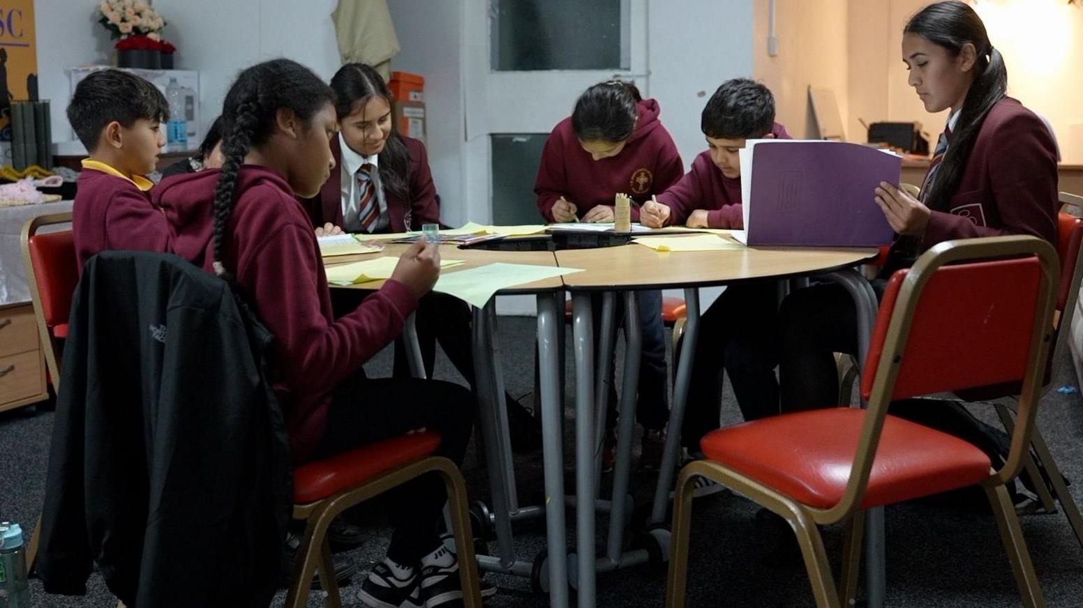 A group of six male and female school pupils of varying ages sitting around a table writing in notebooks and on worksheets. They are all wearing maroon school uniforms. 