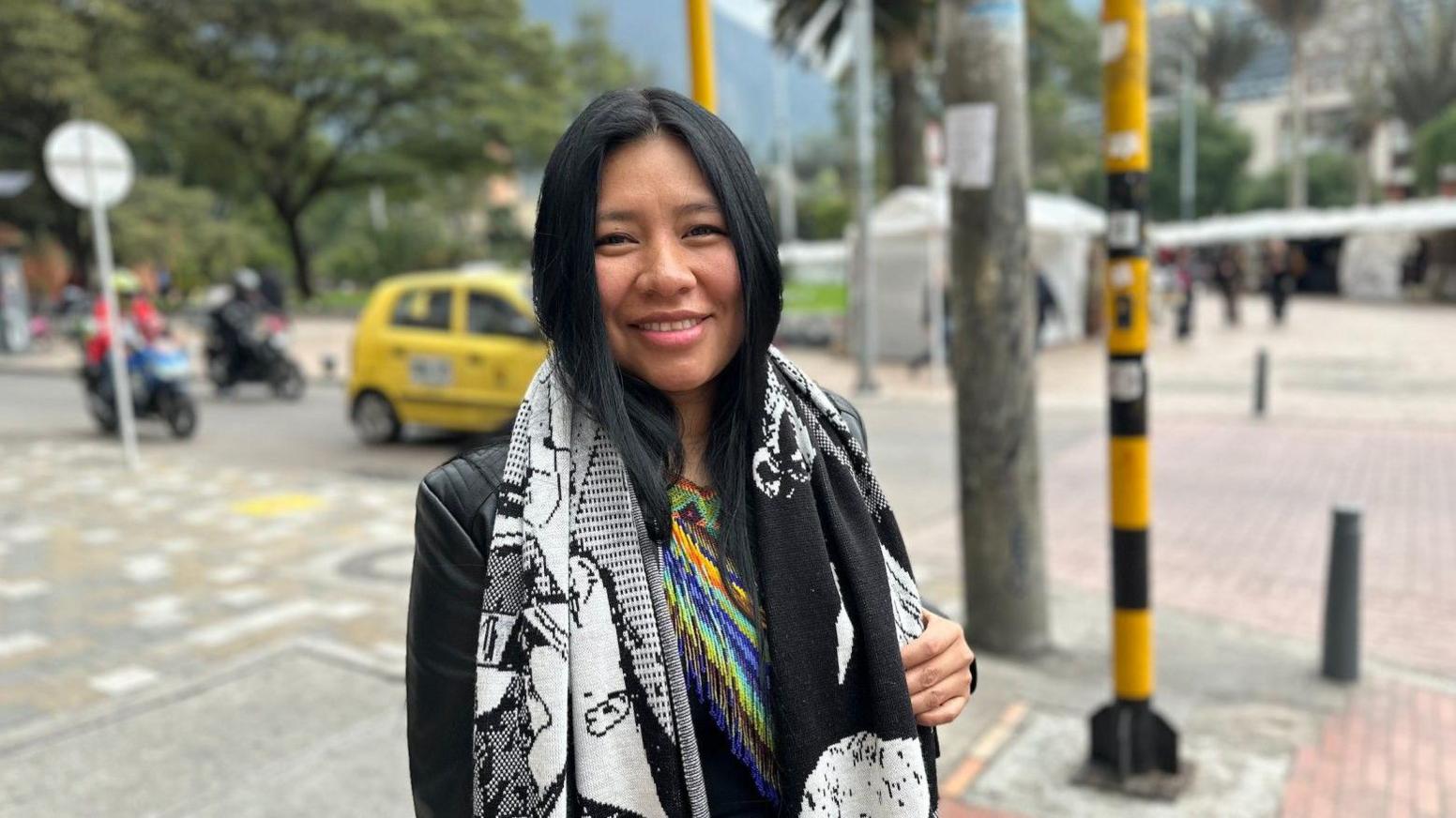 A woman with long dark hair stands at a street intersection with trees, vehicles, and pedestrians visible in the background. She wears a black jacket, a large black-and-white patterned scarf, and a brightly coloured beaded garment. She smiles directly at the camera.
