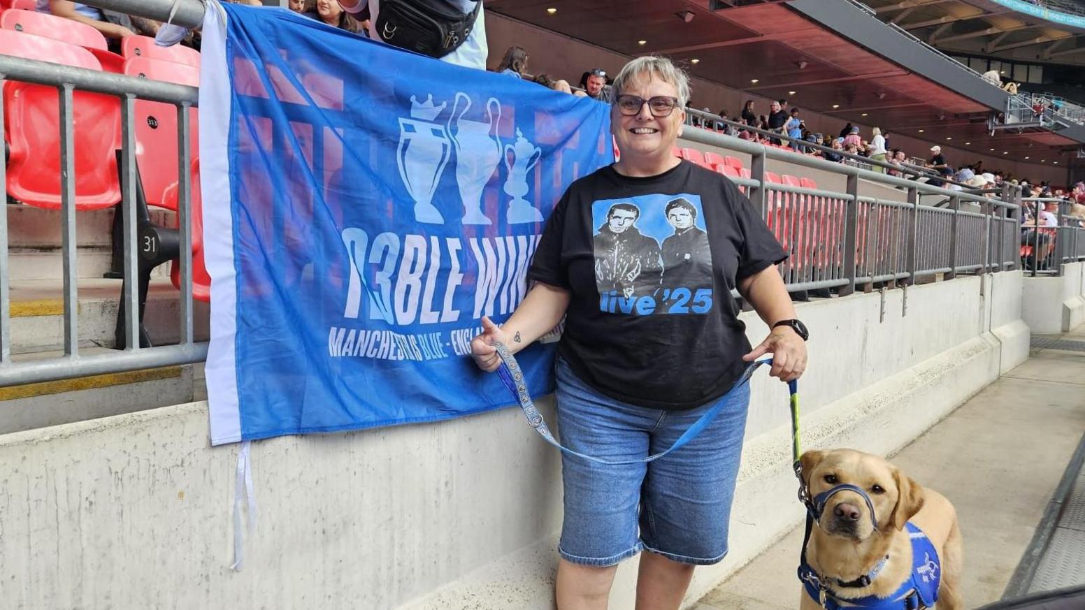 Lynn Ratcliffe wearing long denim shorts and an Oasis T-Shirt, with Barnby, her support Labrador, wearing a blue harness