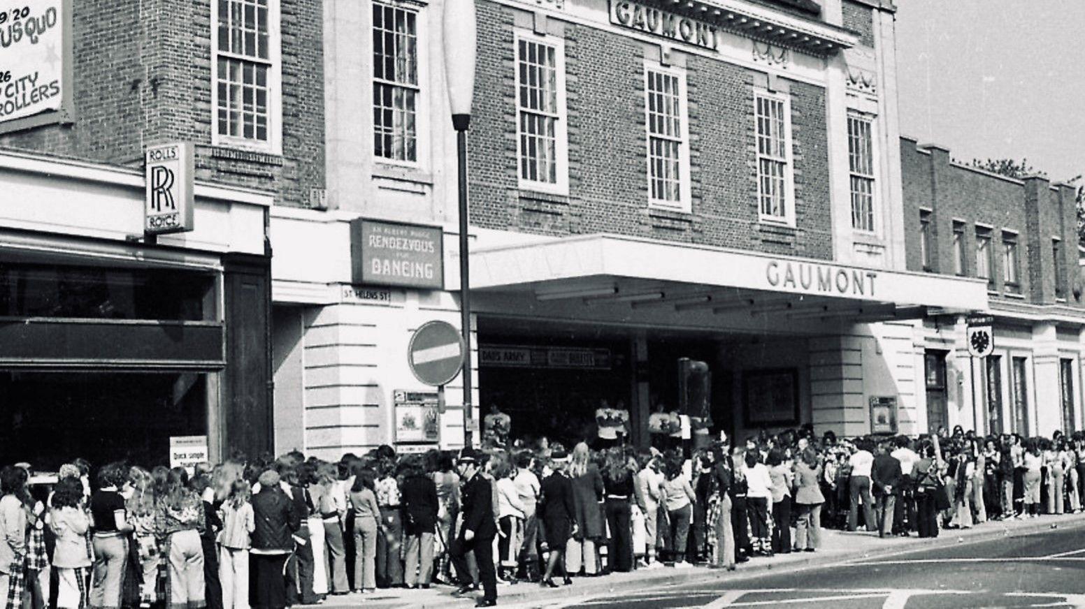 A black and white image taken outside a theatre showing long queues of people outside. The name above the canopy is Gaumont. 