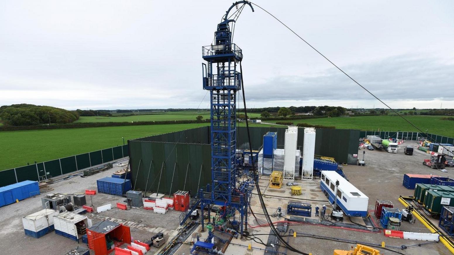 The fracking site is in a fenced off, square area. There is a large blue tower in the middle of a busy area in which there are several large containers, vehicles and equipment. Outside the fenced-off area there are green fields, hedges and trees