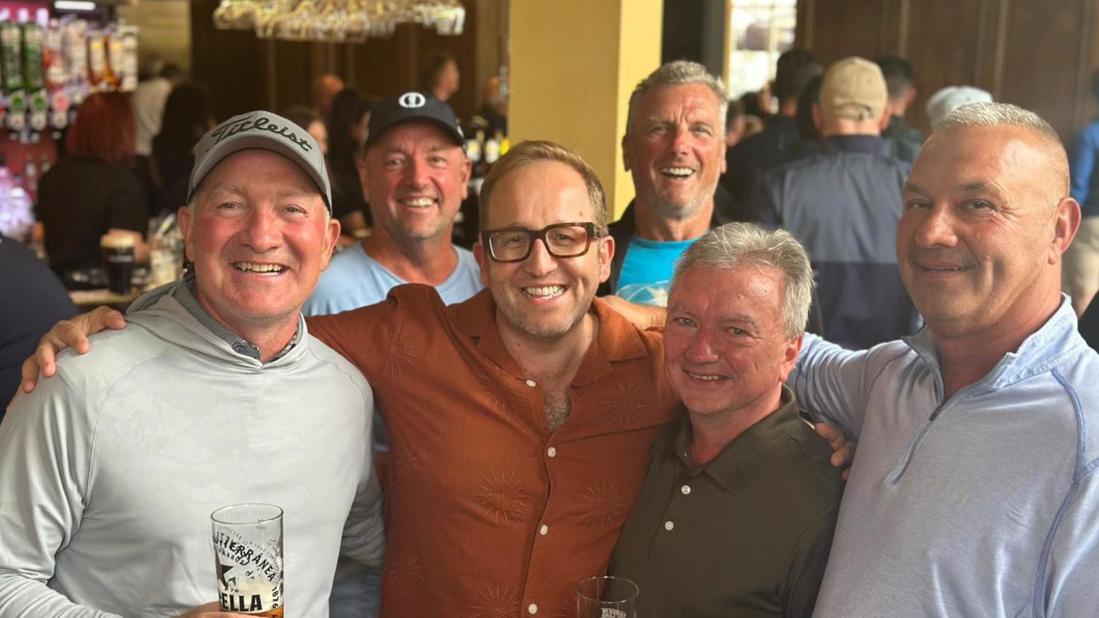 A group of five men have their arms around each others shoulders. There are three at the front and two at the back. The background is a busy bar with loads of people.