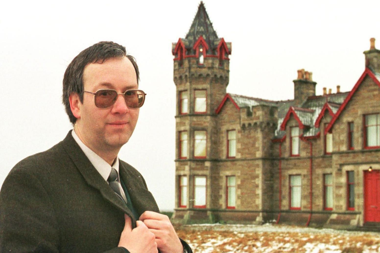 Kevin Booth standing in front of the lodge wearing a suit and tinted glasses and holding his lapels. The building is a large house with a turret.
