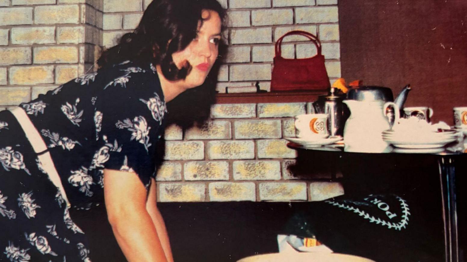 Ruth Jones, when she was 26, with dark wavy hair, red lipstick and in a black and white floral dress. She is on her knees cleaning and looking away from the camera. There is a pile of crockery and teapots on a nearby table and a red handbag on a shelf against a fake brick wall.