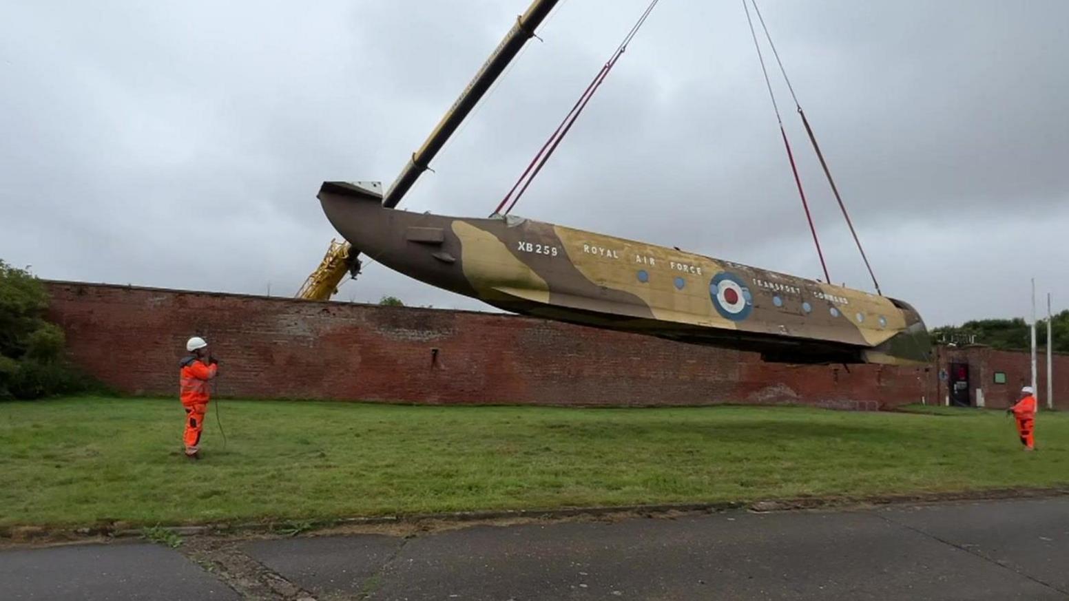 The fuselage of the Blackburn Beverley being lowered over the wall by a crane. Two men are guiding the plane on to grass. 