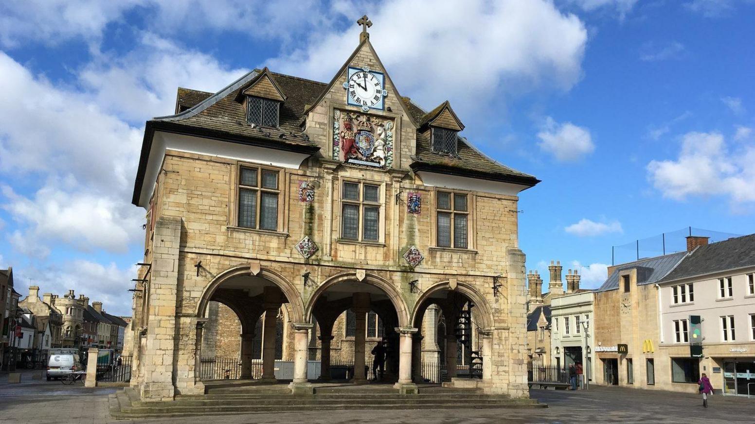The front of Peterborough's Guildhall - a 17th Century stone building with an open area under three arches on a raised plinth at ground level, with two storeys above. There is a a coat of arms and a clock at roof/second storey level.