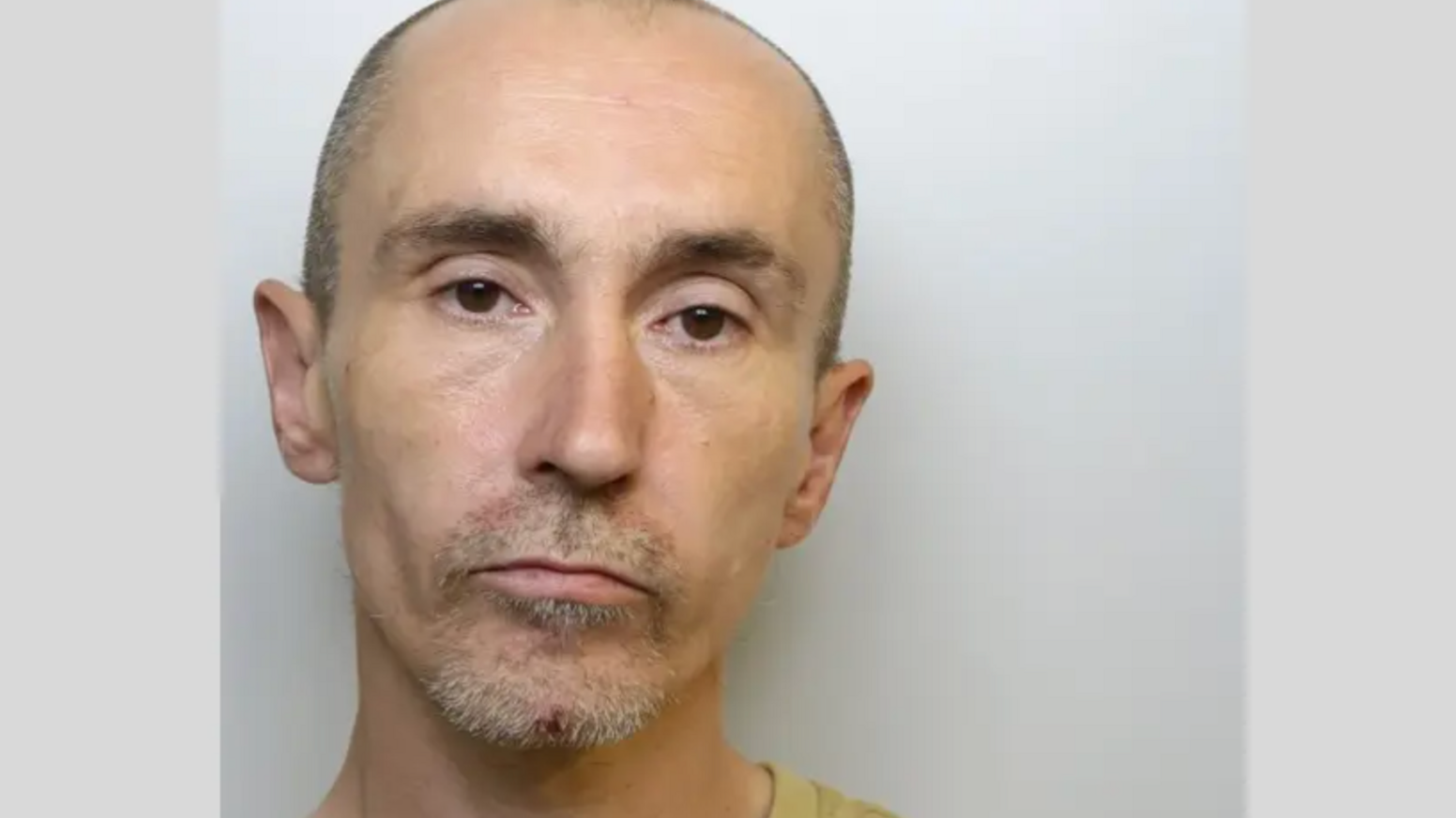 A police custody image of a man in his 40s with a shaved head, stubble and a cut on his chin. He has brown eyed and is wearing a yellow top.