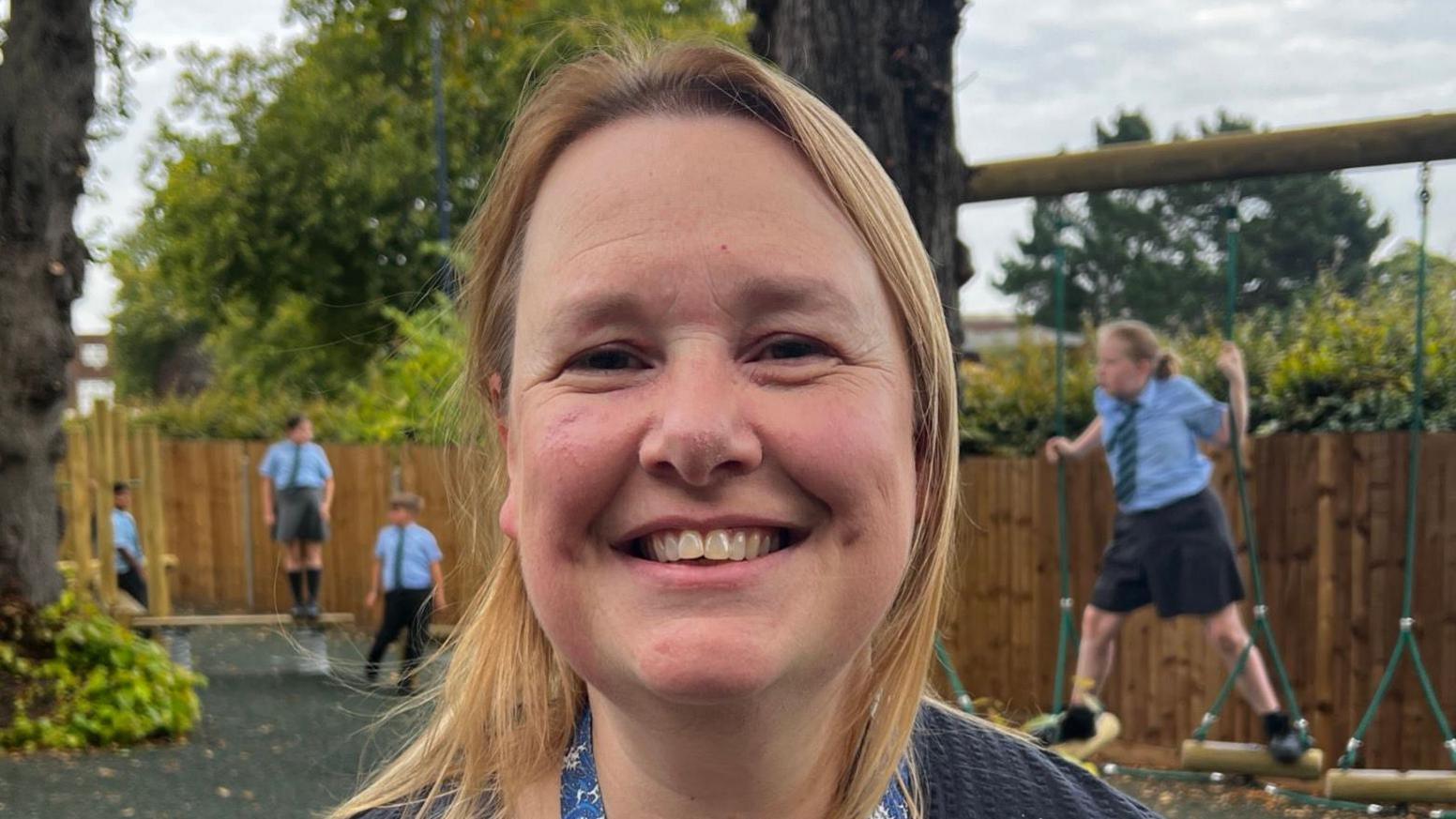 Kirsten Cameron stands in the new play area and smiles at the camera. She has short blonde hair and wears a grey top with a blue lanyard around her neck. Children play on the equipment behind her. 