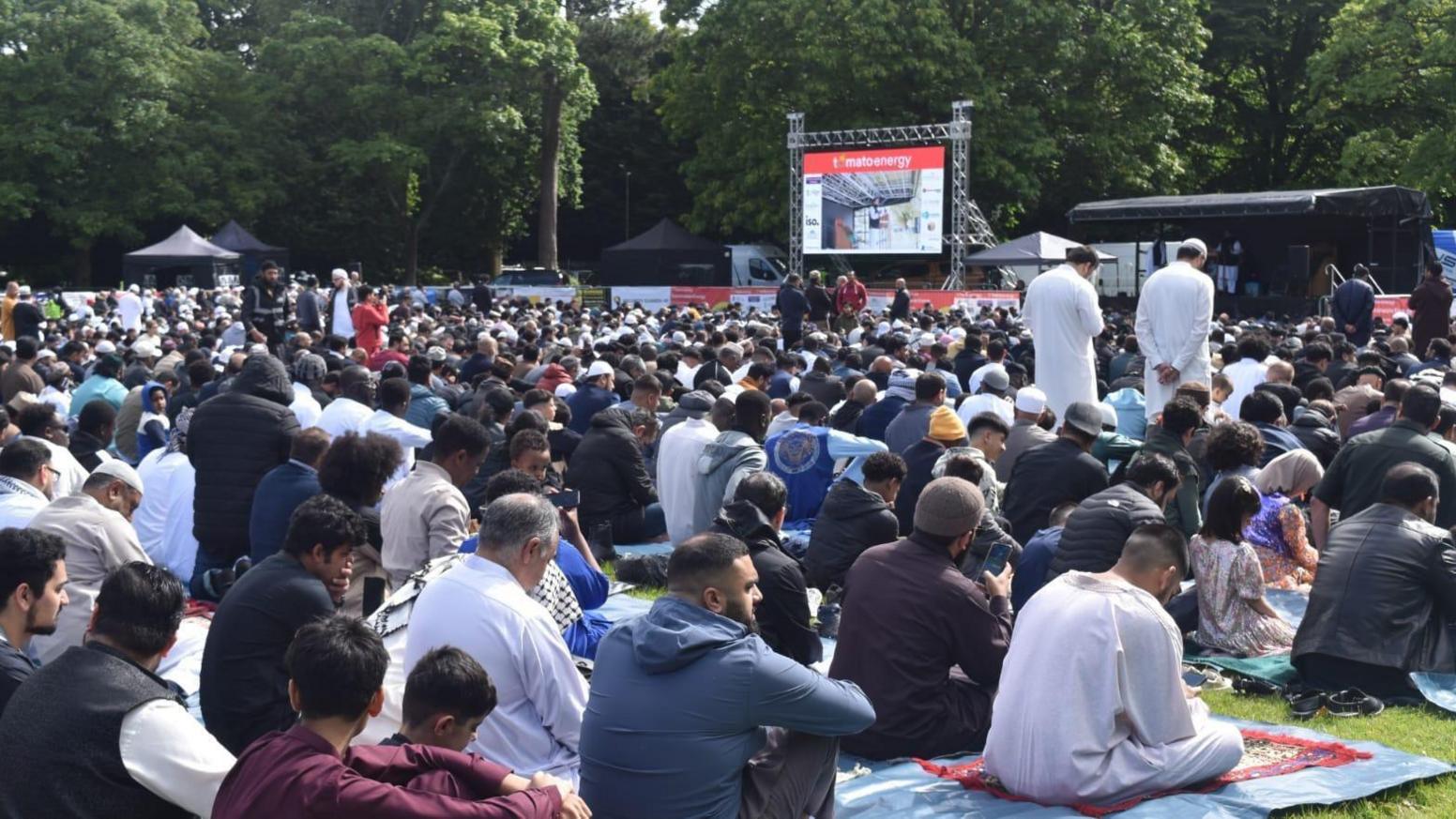 A large crowd of people in a park, most of whom are men and most of whom are sitting, some on prayer mats. In the distance, in front of the crowd, is a large TV screen.