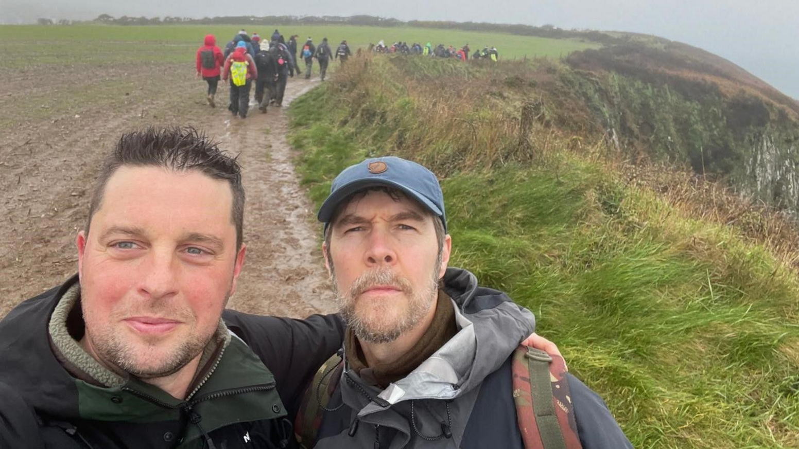 Craig Maxwell smiles and has an arm around comedian Rhod Gilbert's shoulder. The pair are in the middle of a muddy path with a group of hikers in the background.