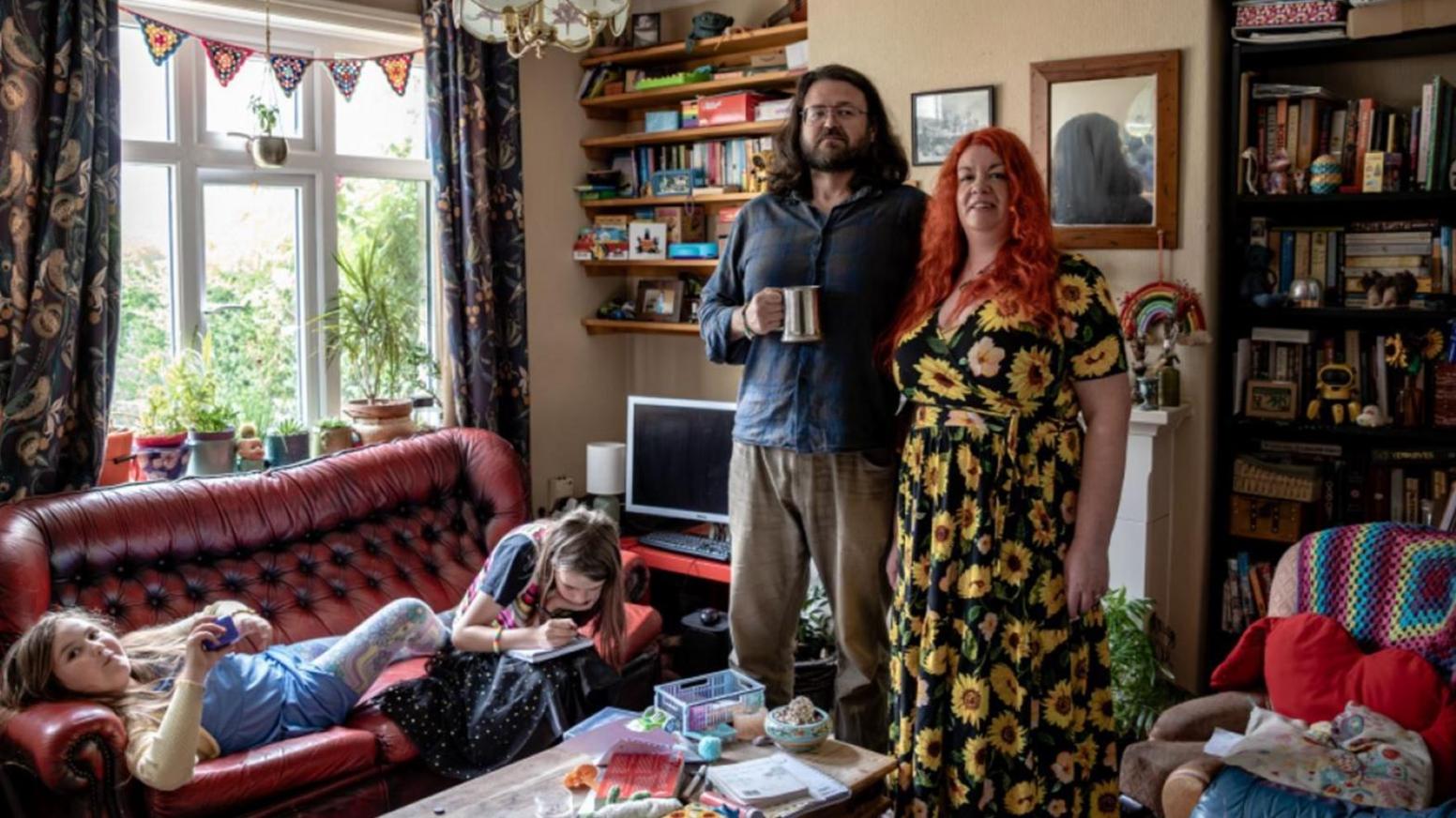 A man and a woman stand in their living room, the woman has red hair and wears a dress with sunflowers on and the man has long brown hair and holds a silver tankard. There are two young girls on a red leather sofa.
