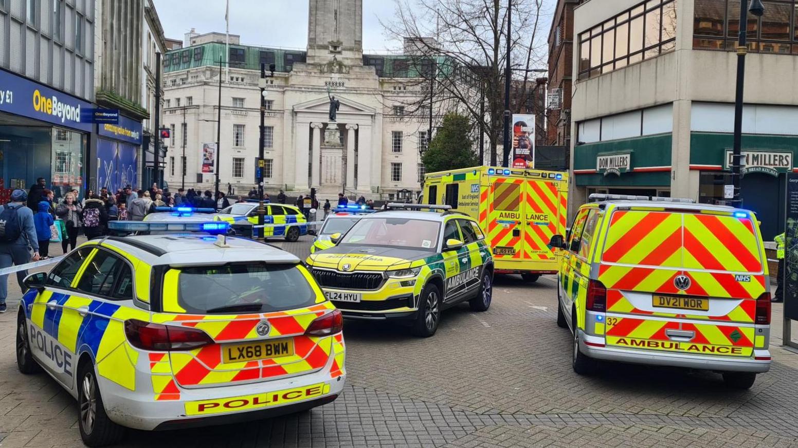Police cars and ambulances parked in the middle of the street in Luton town centre. They are surrounded by blue and white police tape and people milling around.