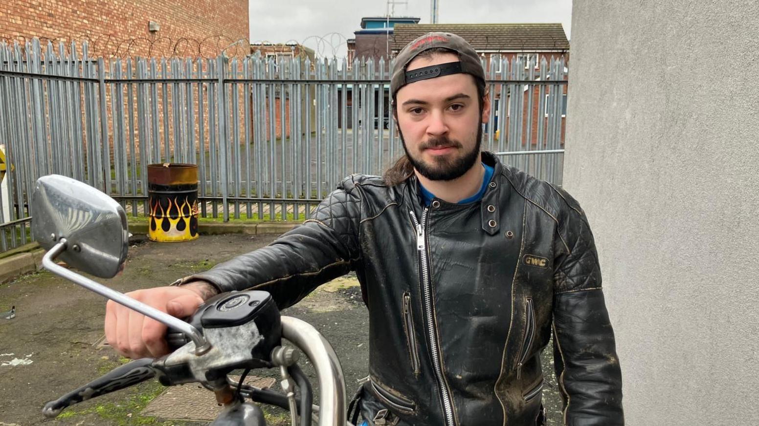 A young man with long brown hair and a brown beard, wears a black leather jacket and a black back-to-front cap, as he sits on a motorbike in a yard.