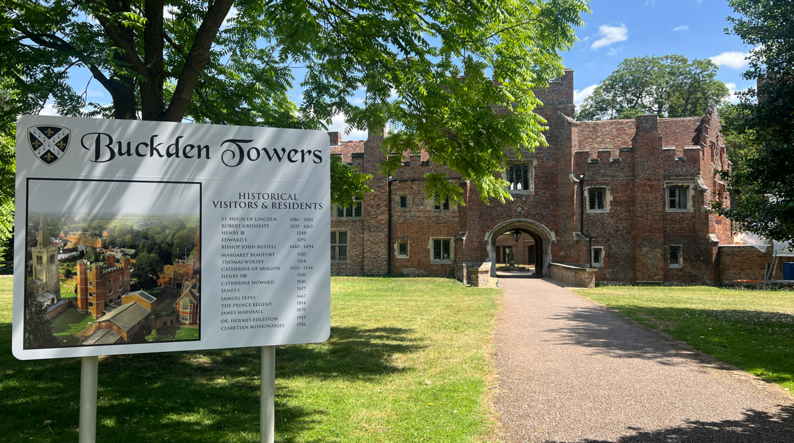 A pathway leading into Buckden Towers. To the left is a sign with information on. The path leads over what looks like a former moat and into an archway where the red bricked towers stand. At either side of it is green grass and some trees stand in the foreground.