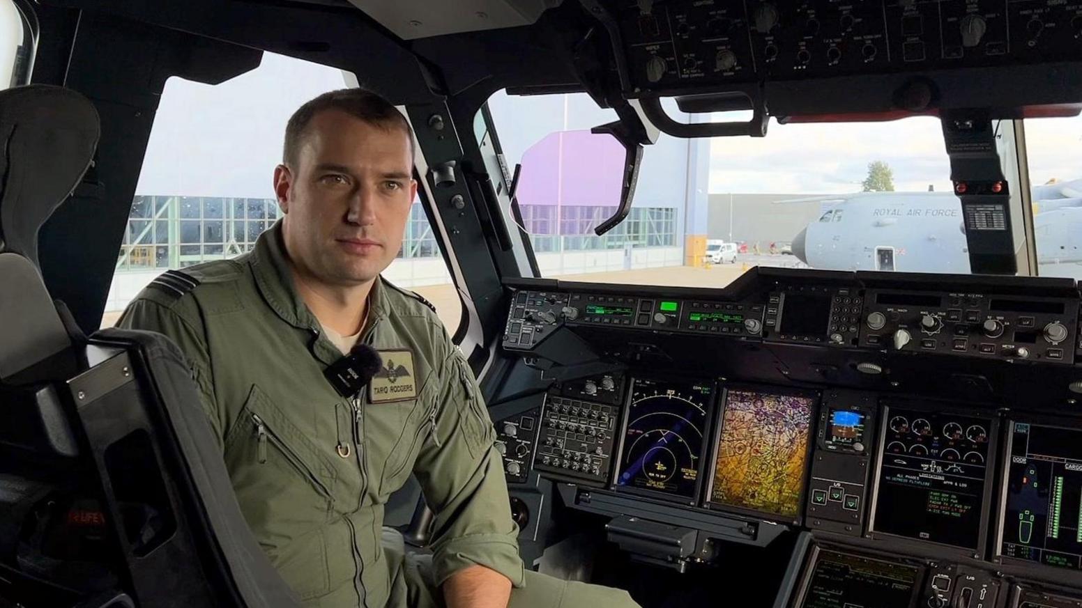 Flight Lieutenant Tariq Rodgers in his flight suit sits in the cockpit of the plane, which has several computer screens alongside the switches and buttons.