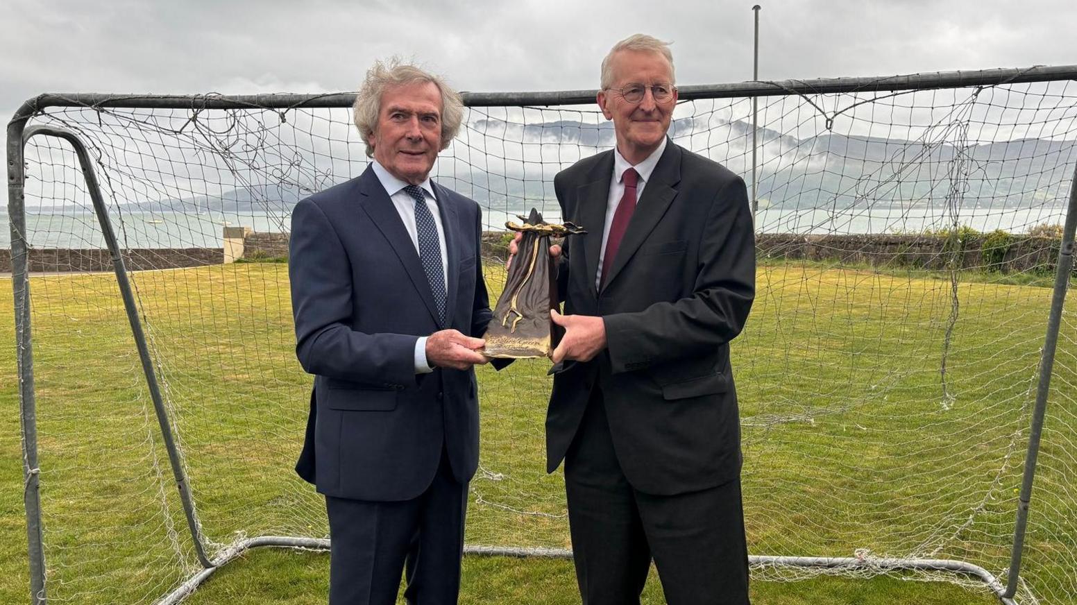Pat Jennings has bushy grey hair and is wearing a suit and is jointly holding a trophy with Hilary Benn in front of a football net