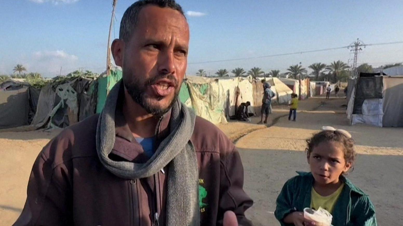 Jemaa Abou Mendi is interviewed for AFP, while a young girl looks on. They air standing on a dusty round in front of a row of makeshift shelters.