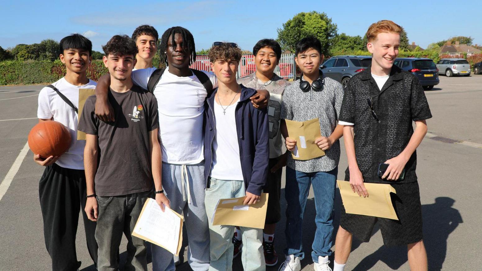 A large group of young men all stand together in their school car park. They are all smiling and looking at the camera, wearing casual attire and holding brown envelopes with their results. In the distance is some green hedging and trees.