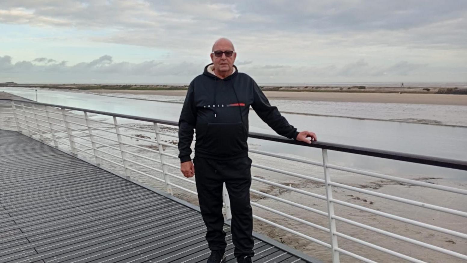 An older man wearing a dark hoodie stands in front of railings overlooking a watery, sandy coastal landscape