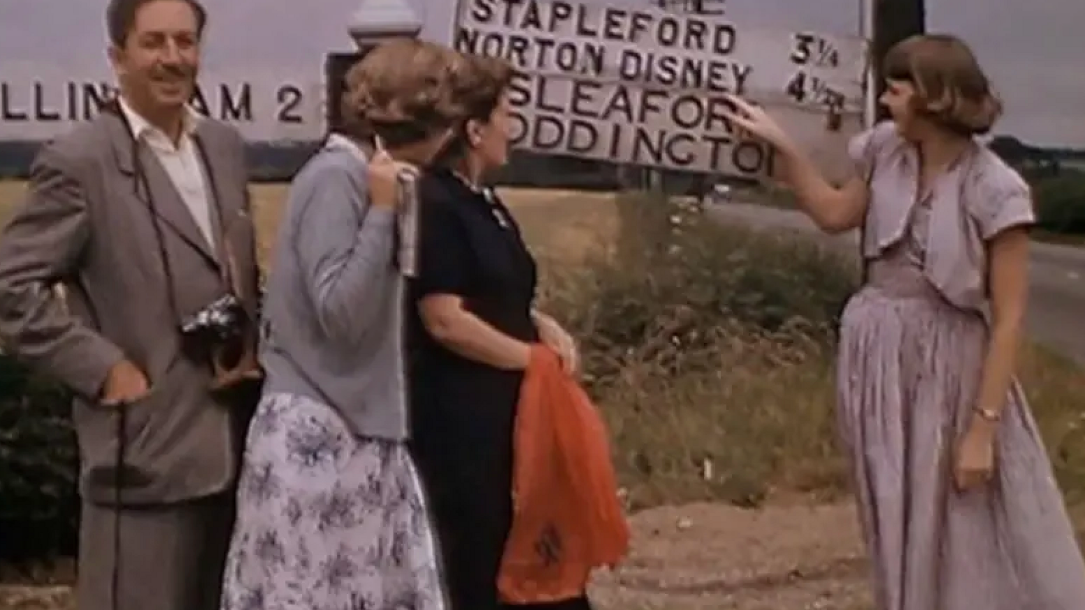 The Disney family are standing in front of a Norton Disney sign when they visited the village in 1949. Walt is on the left in a grey suit and has a camera around his neck, while a young woman in a lilac dress is pointing to Norton Disney on the sign. Two other women are also looking at the sign, which includes Sleaford and Stapleford