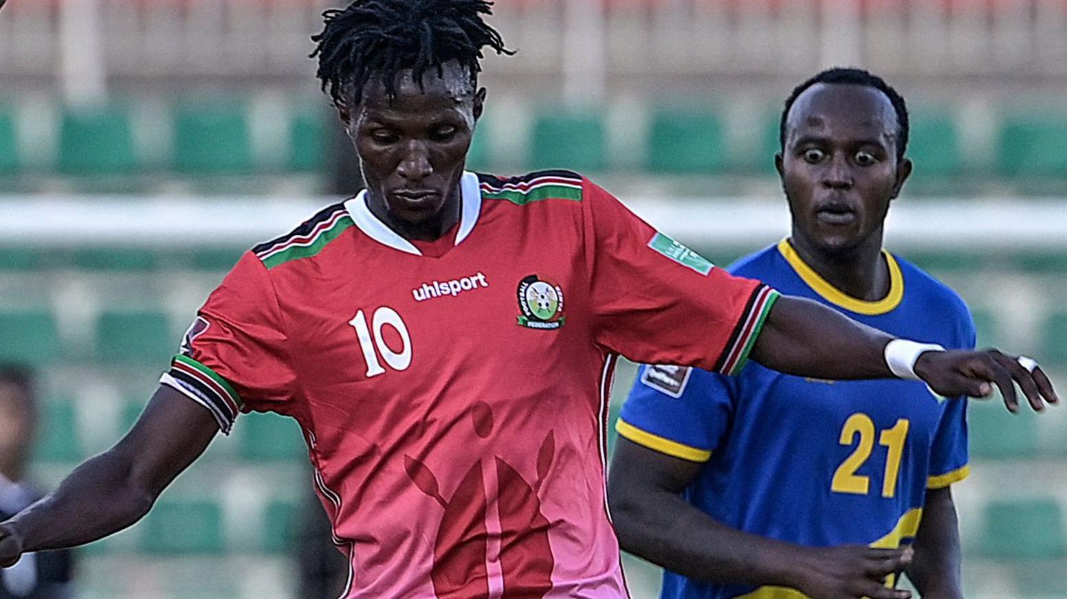 Kenneth Muguna, wearing a red Kenya shirt with the number 10 on the right of his chest, looks down towards his feet during a football match while an opponent wearing blue is pictured in the background