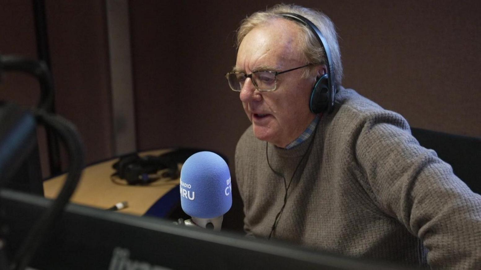 Llun o Dewi Llwyd yn stiwdio radio yng nghanol rhaglen Post Prynhawn. Mae'n gwisgo siwmper frown ac mae ganddo sbectol. Mae'n eistedd o flaen meicroffon glas.