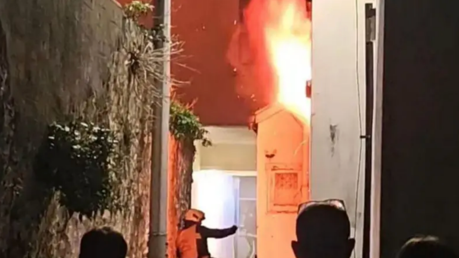 A firefighter inspects the outside of a building which is on fire with flames billowing from the roof. Three people standing at the edge of a cordon can be seen watching on. A large stone wall is to the left of the image.
