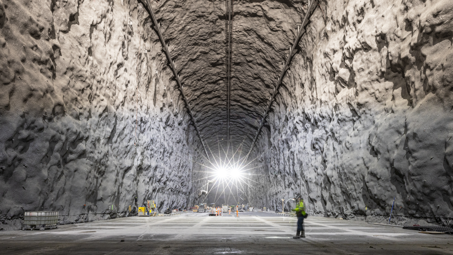 Workers in orange overalls in the background appear like tiny insects in a big white cave, illuminated with bright white electric lights that form two starbursts at the top of the frame. In the foreground a person wearing a green jacket, appearing larger in the frame, studies a laptop.