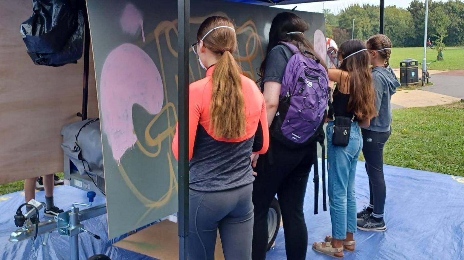 The back views of four teenagers, all with long hair, working on an artwork on a large board in a field, while standing on a blue plastic dustsheet 