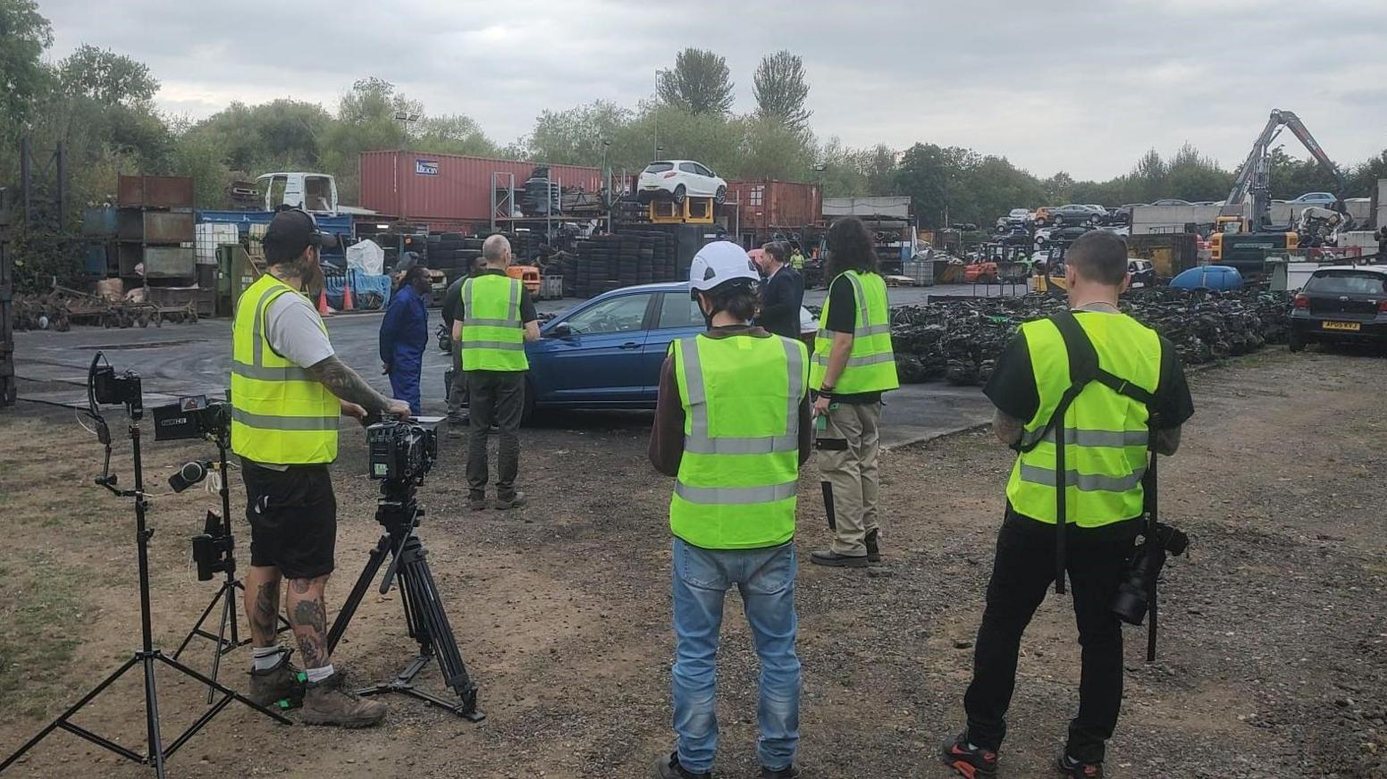 Production crew members wearing hi-vis film scenes at Assington Autos scrapyard in Suffolk. Piles of cars are piled up around them.