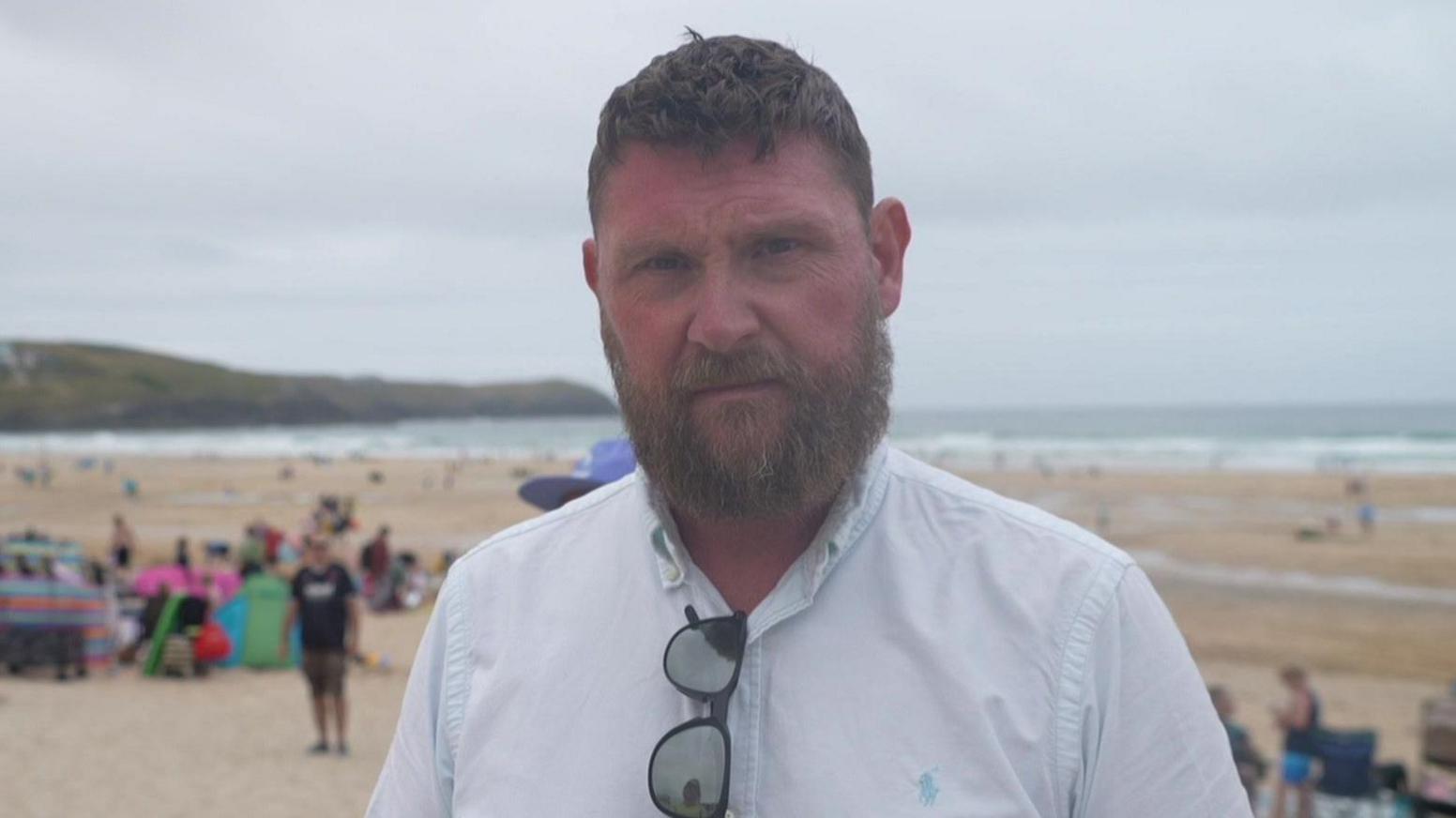 Mayor of Newquay Drew Creek stood on Fistral Beach. He is wearing a white shirt with black sun glasses. There are a number of people on the beach behind him. He is looking straight at the camera and has a big brown beard. 