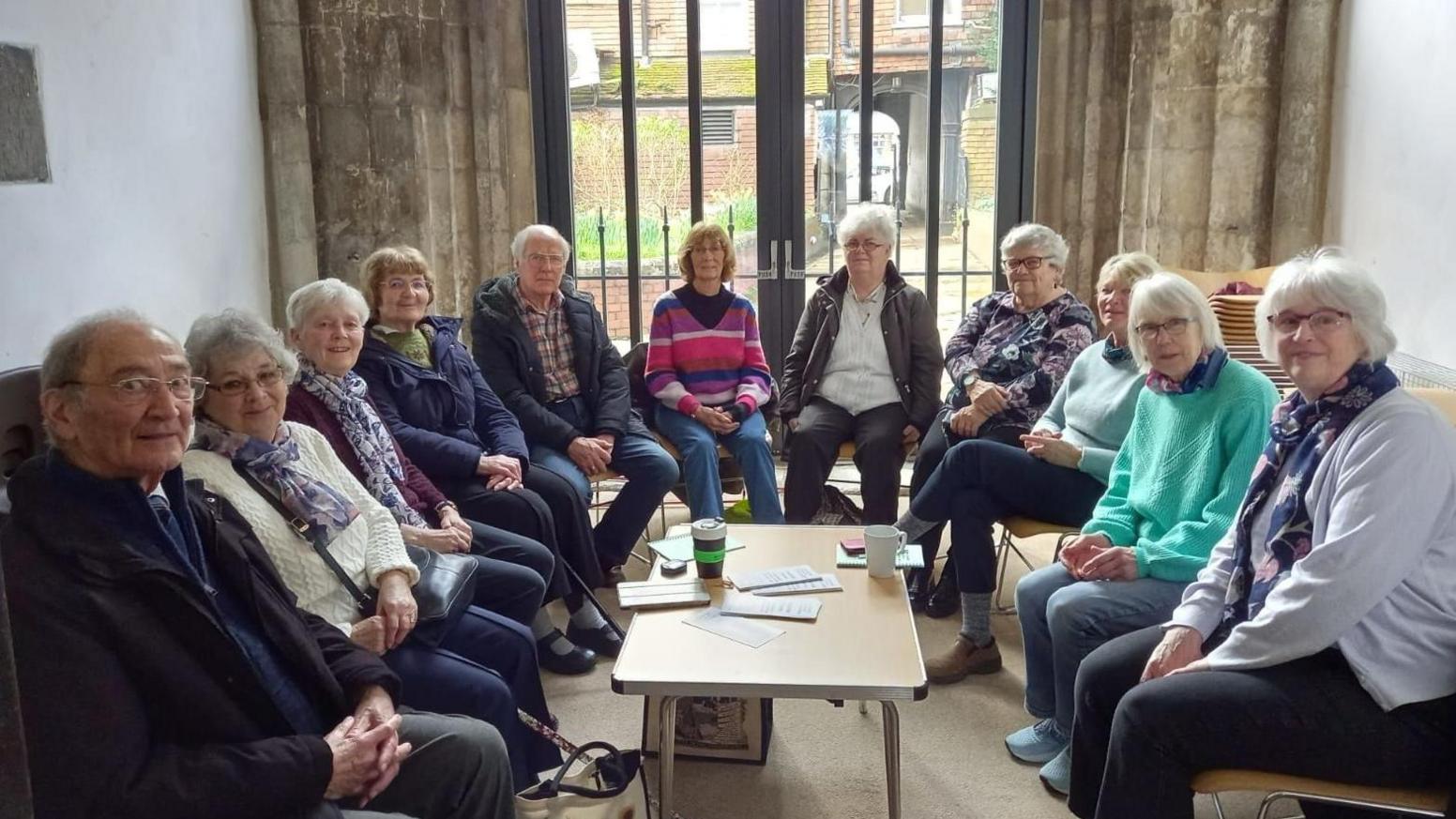Eleven people sat around a little table for the monthly support group. All are looking towards the camera. They are sat in a church, with a window behind them. 