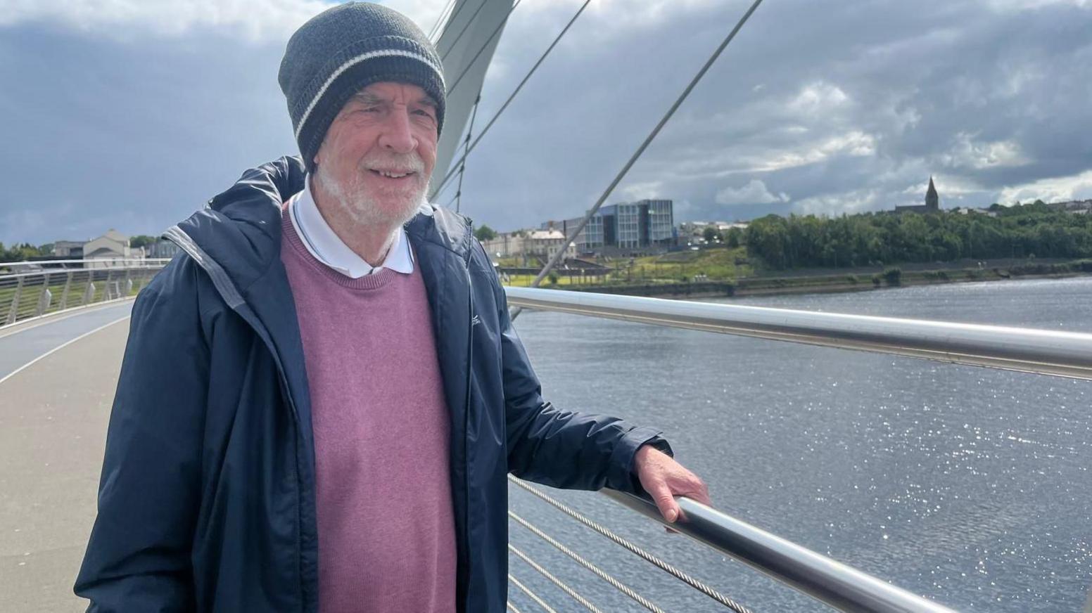 a man stands on a bridge wearing a blue jacket and pink jumper. he has a grey hat on his head. a river is behind him, and a number of buildings can be seen in the background