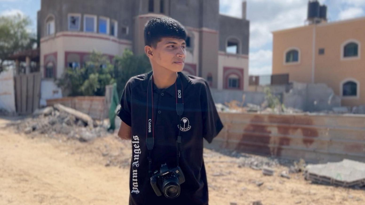 Diya al-Adini stands in an empty Gaza street, with both arms missing and one stump visible underneath a black t-shirt and a camera hanging around his neck