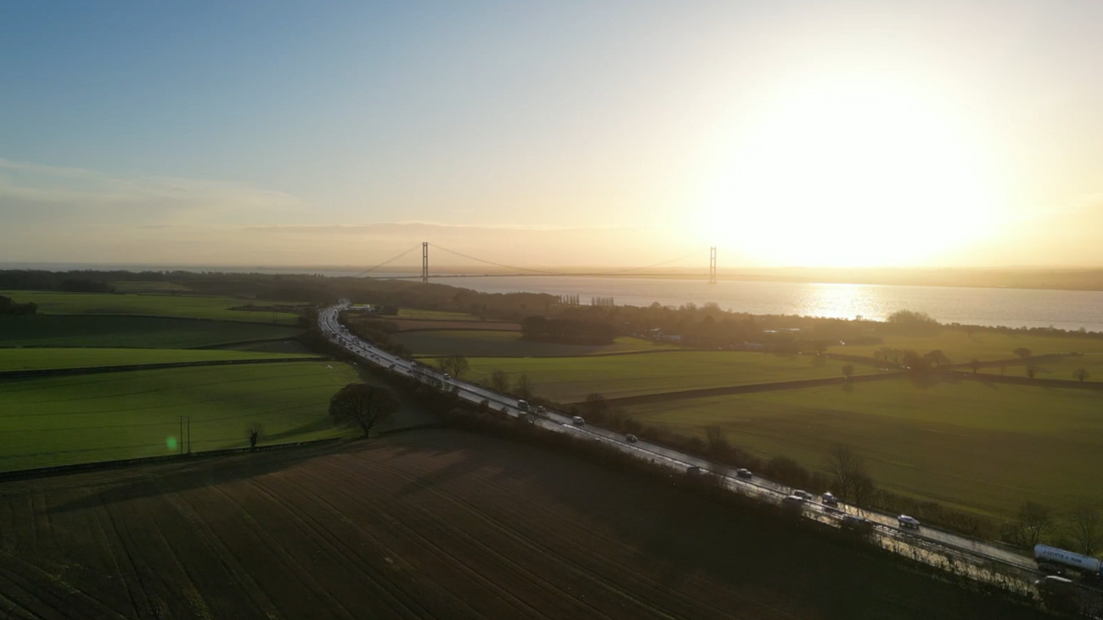 A drone image of East Yorkshire. The sun is rising over the Humber Bridge which visible in the distance along with the Humber Estuary. Cars and lorries are visible on the A63 road which is running across the image and flanked by green fields.