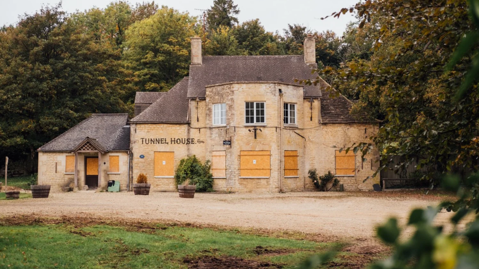 A photograph of a traditional sandstone building with two chimneys jutting out. The lower windows are boarded up with plywood. Traditional lettering on the exterior reads: "Tunnel House".